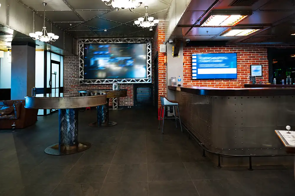 Modern bar interior with metal and brick decor, high tables, bar counter with stools, large screen, and digital menu displays.