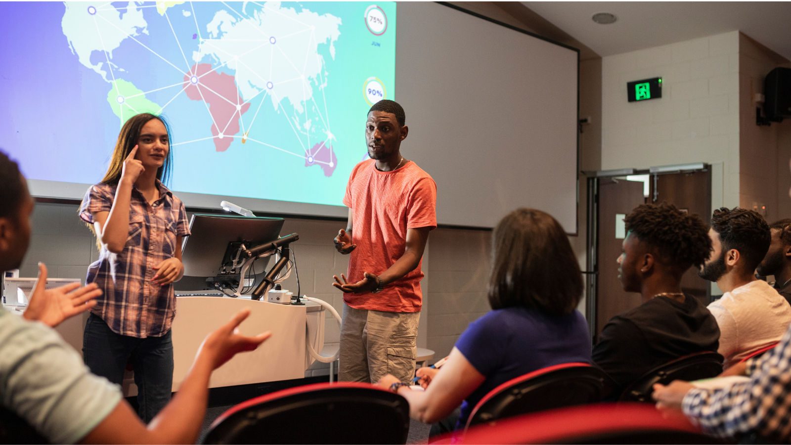 Two presenters stand at the front of a classroom speaking to an audience, with a world map projected on a screen behind them.