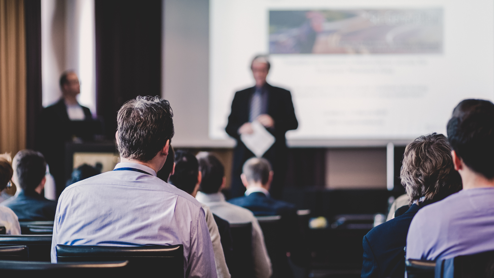 People seated in a conference room listen to two presenters speaking at the front near a large screen displaying a presentation.
