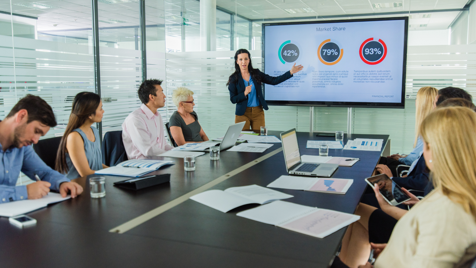 A woman stands at the front of a conference room giving a presentation to colleagues, with a screen displaying market share percentages behind her.