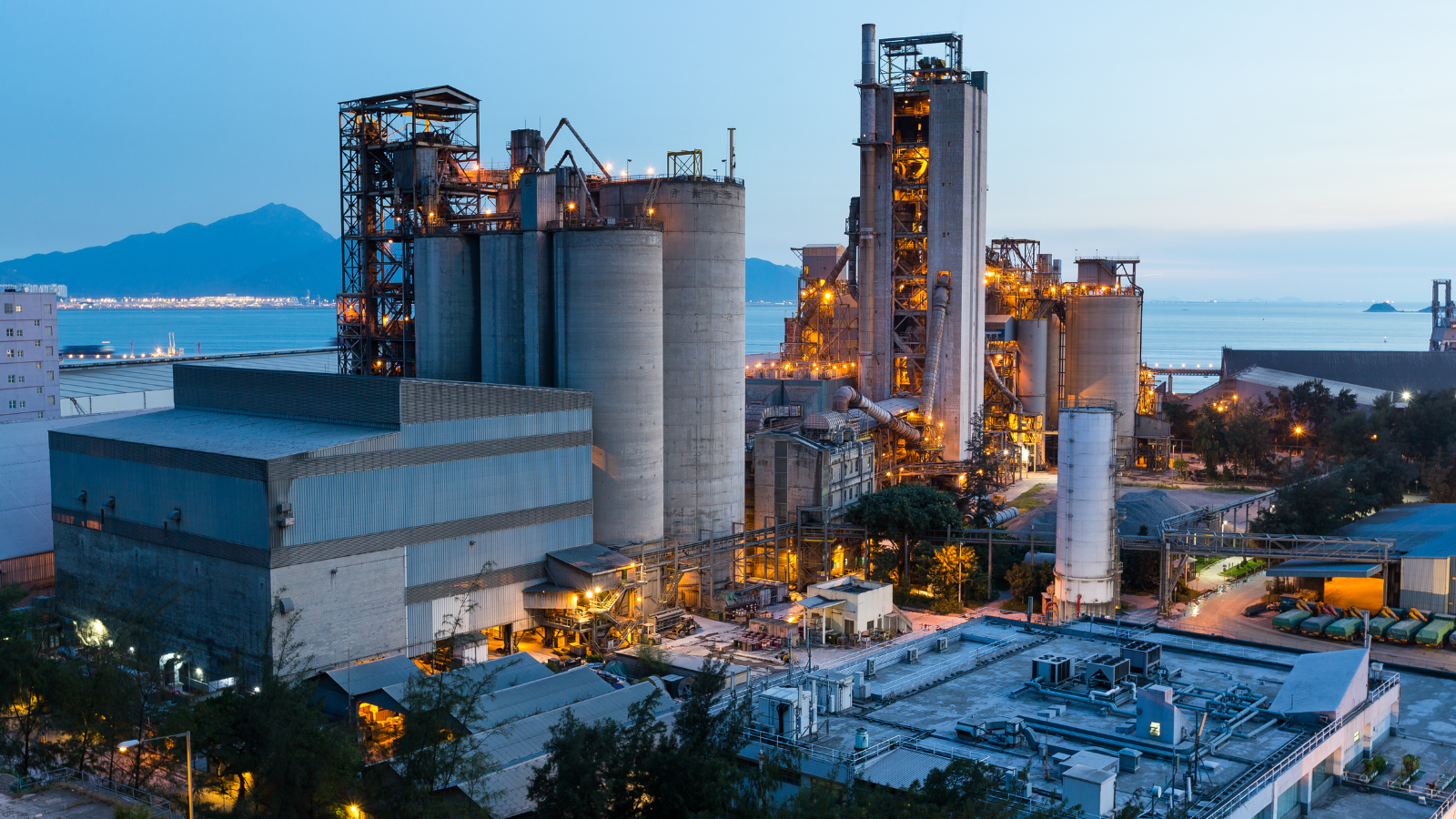 An industrial cement plant with large silos and towers is illuminated at dusk, with mountains and water visible in the background.