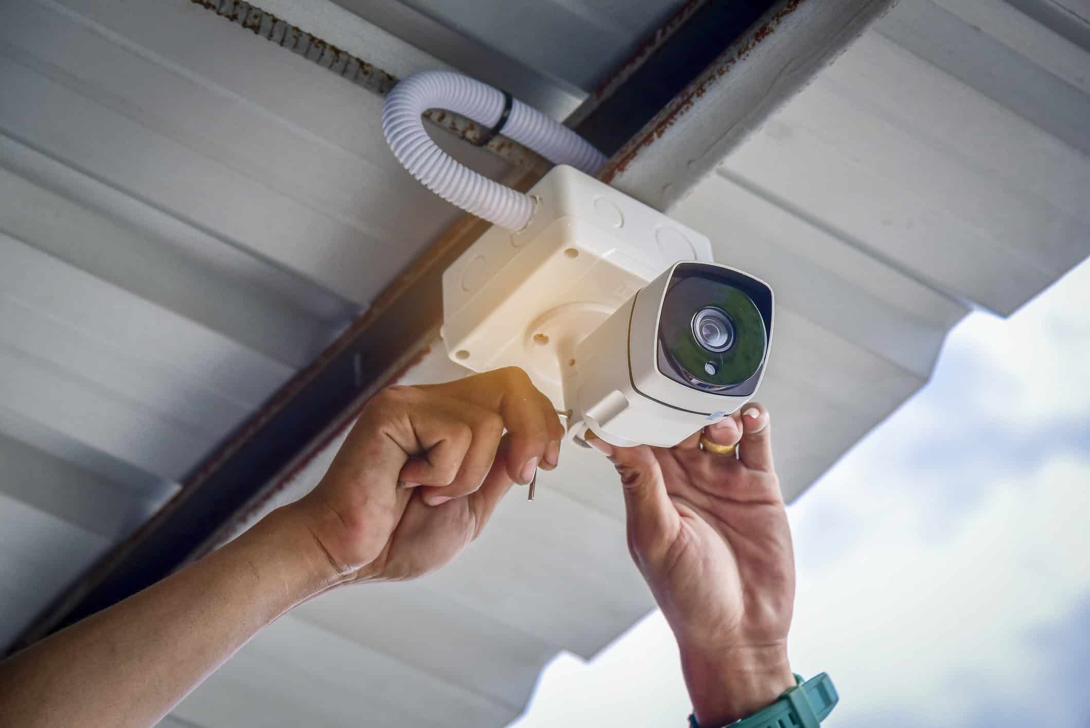 A person installs a security camera on a metal ceiling using a screwdriver.