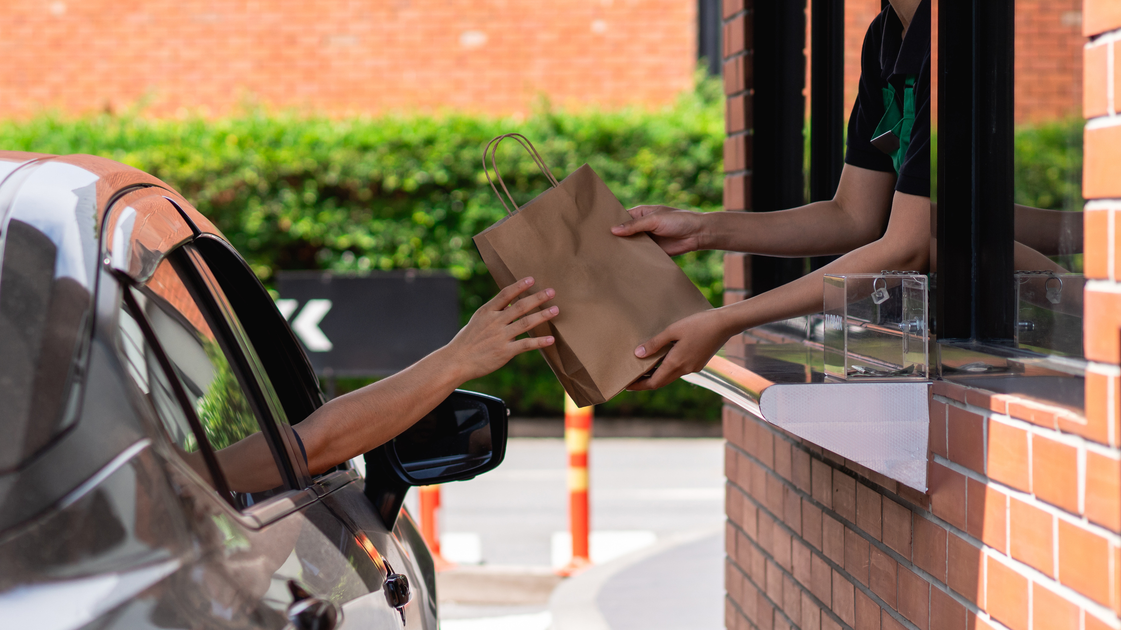 Drive -thru technology maintain and install CCTV and audio visual A person in a car receives a brown paper bag from an employee at a drive-thru window, with brick walls and greenery in the background.
