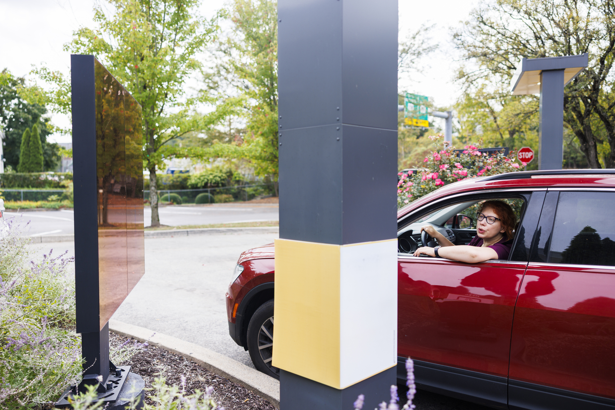 Drive thru tech install and maintain  for quick service restaurants. A person in a red SUV waits at a drive-thru ordering station, looking toward the menu. Trees and flowers are visible in the background.