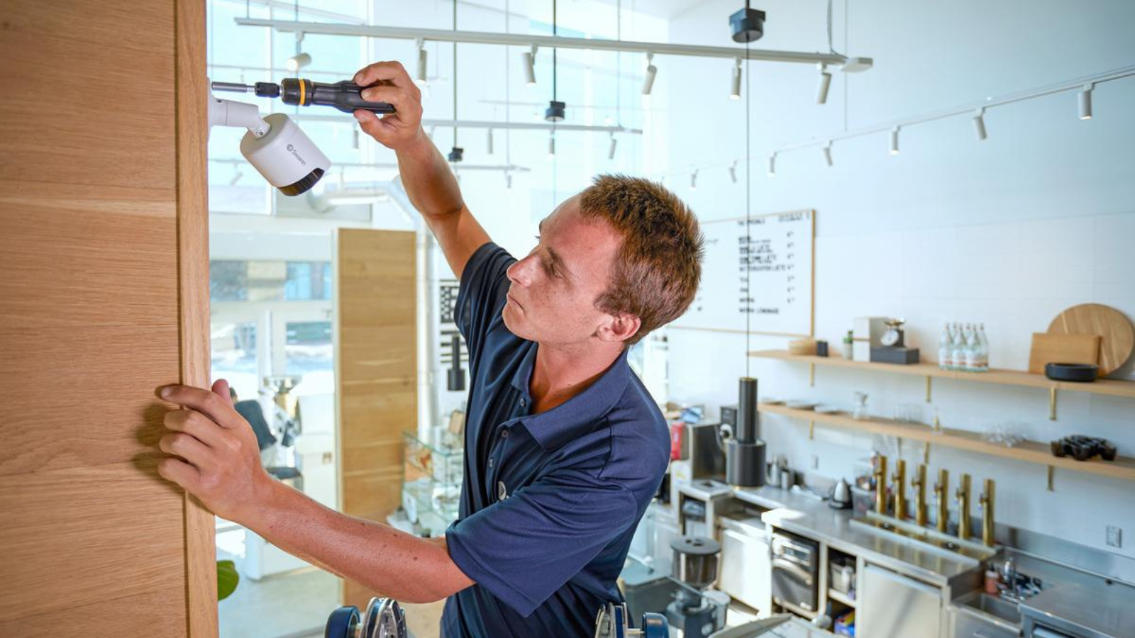 A man in a blue shirt installs a security camera on a wooden wall inside a modern kitchen or QSR restaurant, complete with sleek metal appliances and shelves&mdash;an environment that could benefit from both enhanced security and digital menu board upgrade solutions.