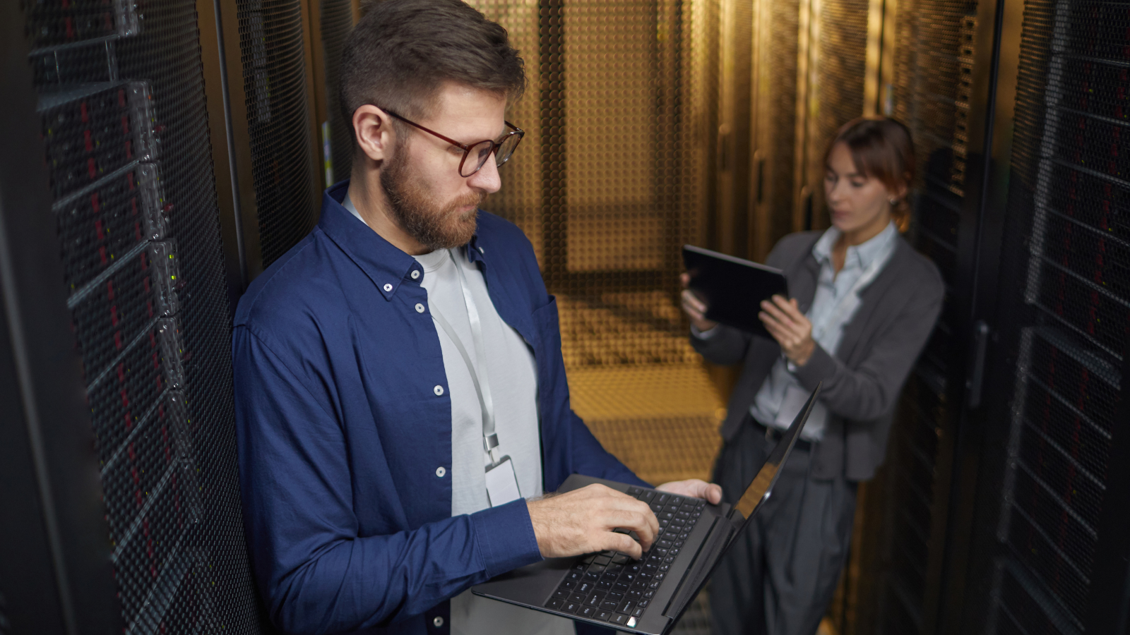A man using a laptop in a server room focuses intently, aware of the NRBES 2027 deadline approaching in Q4 2026, while a woman in the background&mdash;possibly a McDonald&rsquo;s franchisee&mdash;works on a tablet among server racks.